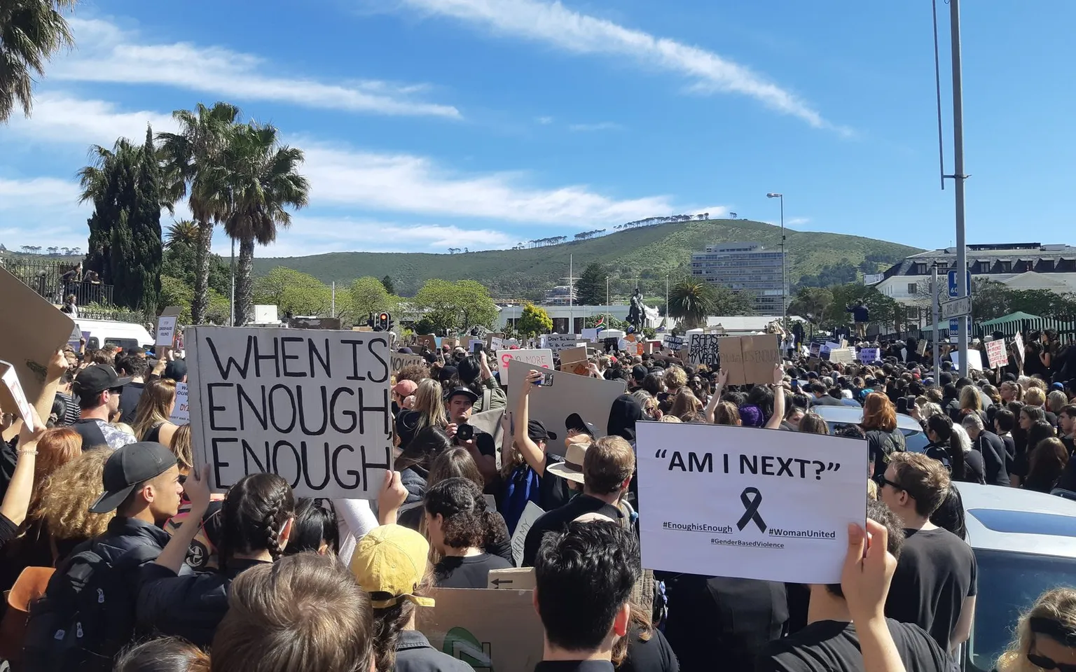 Thousands of protesters fill a Cape Town street outside Parliament. Two hand-made placards are raised above the crowd: "When is enough, enough" and "Am I next?"